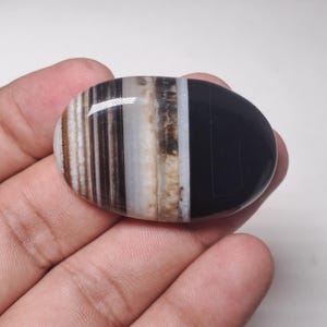 May include: A polished, oval-shaped gemstone with a mix of colors. The stone features bands of brown, white, and black, with a solid black section on one side. The stone is held in a hand.