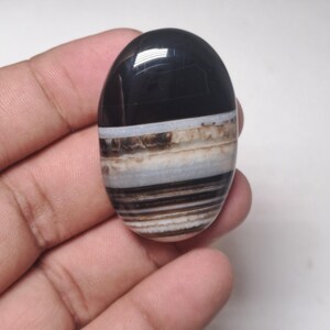 May include: An oval-shaped polished stone with a black top and layered bands of white, brown, and tan. The stone is held in a hand, showcasing its smooth, glossy surface. The gemstone has a natural, earthy appearance.