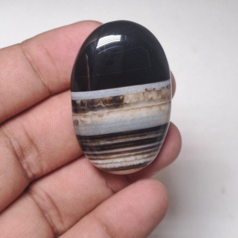 May include: An oval-shaped polished stone with a black top and layered bands of white, brown, and tan. The stone is held in a hand, showcasing its smooth, glossy surface. The gemstone has a natural, earthy appearance.