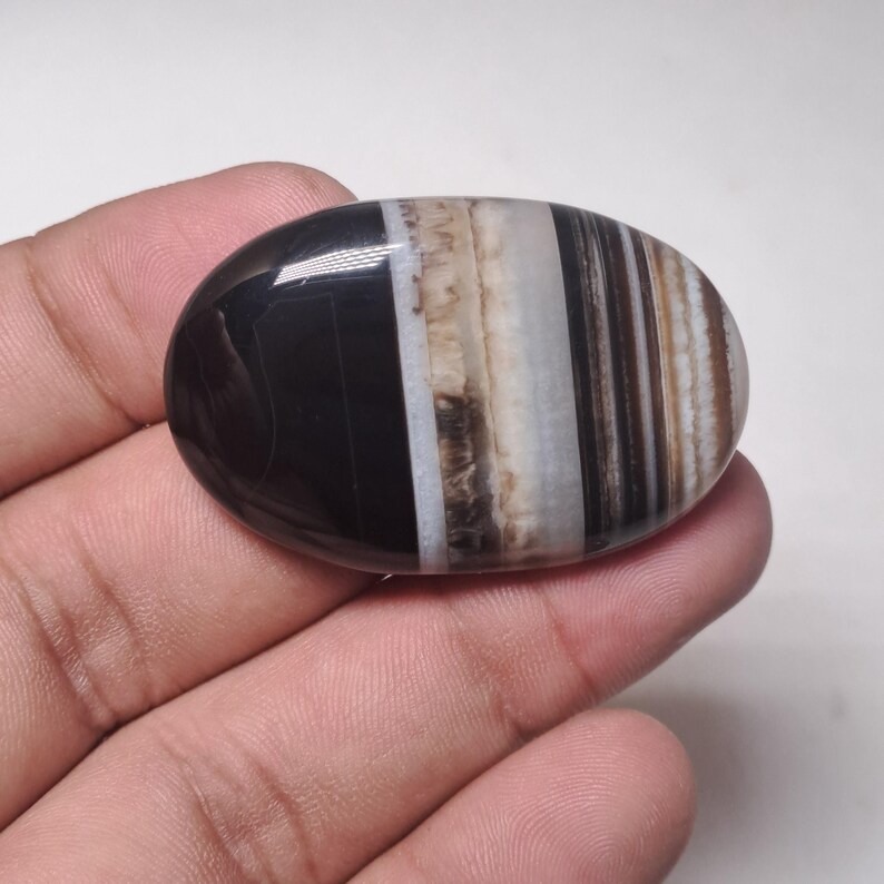 May include: A polished, oval-shaped gemstone with a striking pattern of black, white, and brown bands. The stone is held in a hand, showcasing its smooth, glossy surface. The bands create a striped effect, adding visual interest to the natural stone.