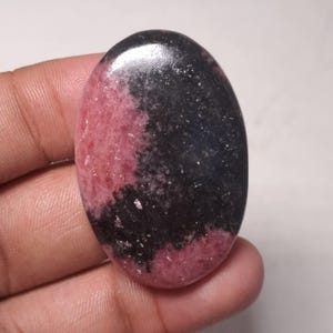 May include: A smooth, oval-shaped cabochon gemstone with a black and pink pattern. The stone has a polished surface and a speckled appearance.