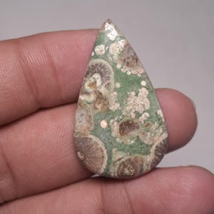 Puede incluir: Un cabujón de ágata verde, marrón y blanco con forma de lágrima. La piedra tiene un patrón de colores y texturas en espiral.