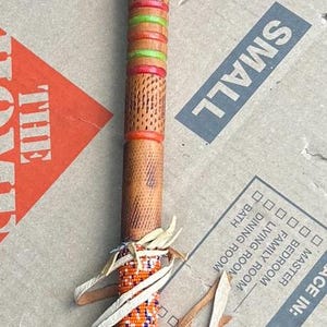 May include: A wooden pipe with a T-shaped copper bowl at the top. The stem features textured wood, horizontal bands of orange and green paint, and a section wrapped in orange, white, and blue seed beads with dangling light-coloured leather fringe.