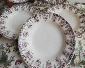May include: Five white ceramic plates with a brown floral pattern around the rims. The plates are arranged on a patterned tablecloth and are of varying sizes. The floral design is detailed and adds a vintage aesthetic.