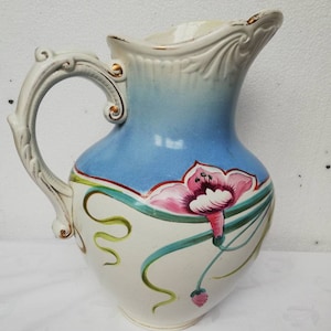 May include: A white ceramic pitcher with a blue and white ombre glaze. The pitcher has a floral design with pink flowers and green leaves. The handle is ornate and has gold accents.