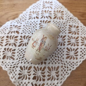 Könnte beinhalten: Eine kleine, cremefarbene Keramikvase mit glänzender Oberfläche und engem Hals. Auf der Vorderseite befindet sich eine detaillierte Illustration von zwei Figuren in traditioneller Kleidung auf einem Feld, ergänzt durch Text und ein Logo.