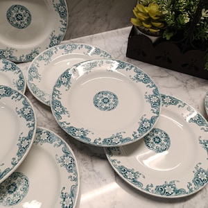 Peut inclure: Un ensemble de six assiettes plates en céramique blanche avec un motif floral vert autour du bord. Les assiettes sont empilées sur une surface en marbre.