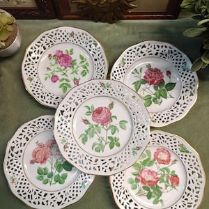 Puede incluir: Cinco platos de porcelana blanca con intrincados bordes calados y ribete dorado. Cada plato presenta un diseño floral diferente con rosas rosas y hojas verdes. Los platos están dispuestos sobre una superficie verde.