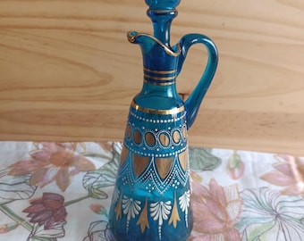May include: A turquoise glass decanter with a handle and a stopper. The decanter is adorned with gold and white decorative patterns. The design includes gold bands and detailed floral and dot patterns. The decanter sits on a floral patterned cloth.