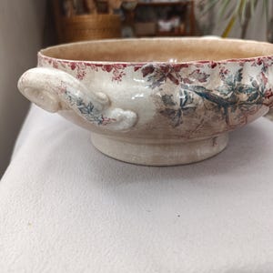 May include: A ceramic bowl with a cream-colored base and a red and blue floral transferware pattern. The bowl features two side handles, a scalloped rim, and a pedestal base. The surface has a visible crackle glaze finish throughout the exterior.