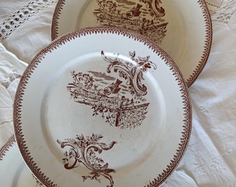 May include: White ceramic plates with a brown decorative border and a detailed brown landscape design. The plates have a vintage aesthetic and are arranged on a white lace tablecloth. The plates are part of a dinnerware set.