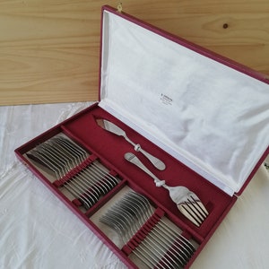 Peut inclure: Un service de couverts vintage dans un étui doublé de rouge. L'ensemble comprend plusieurs fourchettes, couteaux et cuillères, tous avec une finition argentée. L'étui est ouvert, révélant les ustensiles soigneusement rangés.