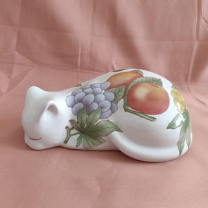 May include: A white ceramic cat figurine in a resting pose, decorated with colorful fruit illustrations. The cat's body features images of grapes, peaches, and other fruits with green leaves. The figurine is on a pink background.