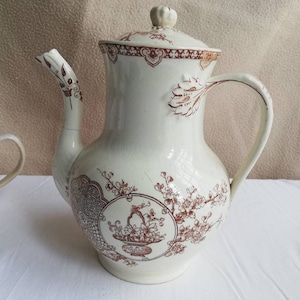 Peut inclure: Une théière en céramique blanche avec un motif floral brun. La théière a un couvercle et une poignée. Le motif représente un panier de fleurs et un motif répétitif de feuilles et de fleurs.