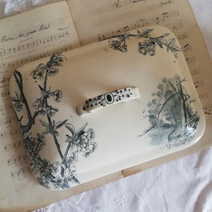 Op de afbeelding: Een wit keramiek deksel met een zwart-wit bloemenontwerp. Het deksel heeft een zwarte handgreep en ligt op een blad muziek.