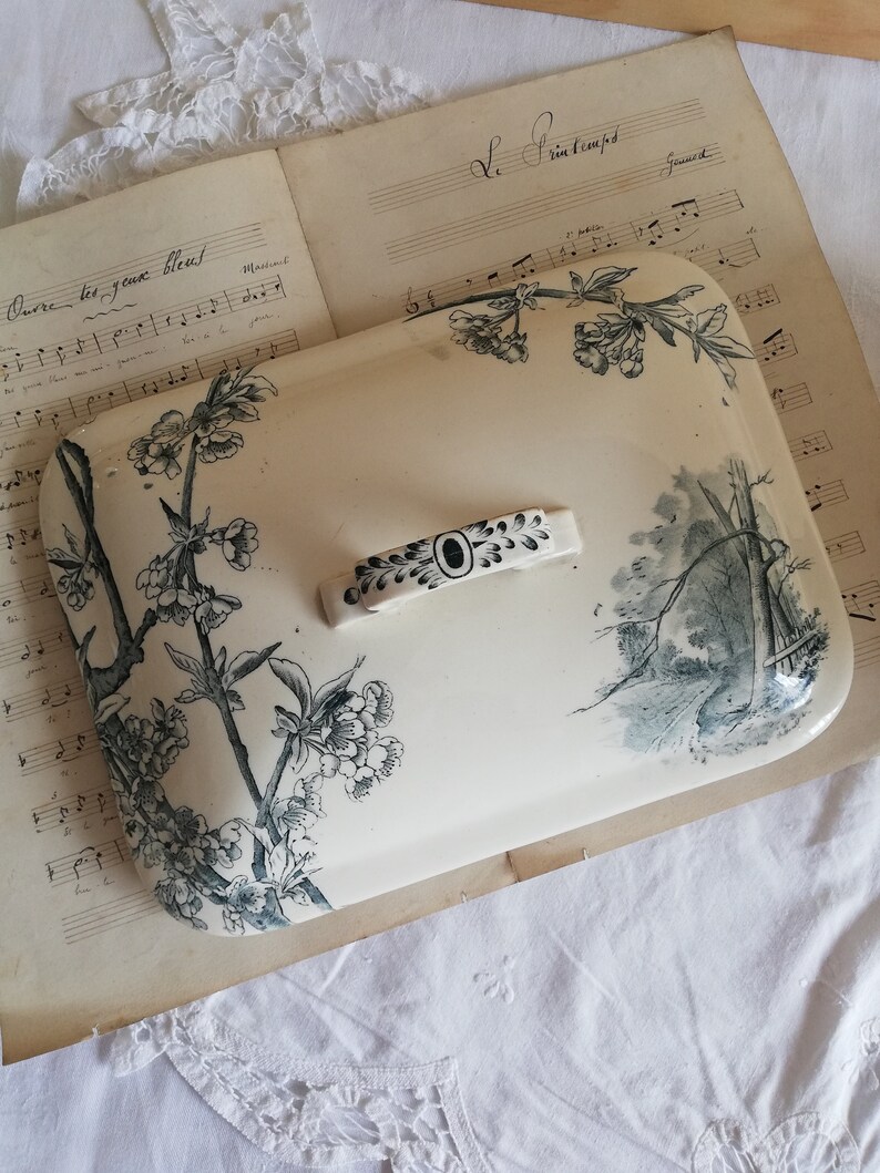 Op de afbeelding: Een wit keramiek deksel met een zwart-wit bloemenontwerp. Het deksel heeft een zwarte handgreep en ligt op een blad muziek.
