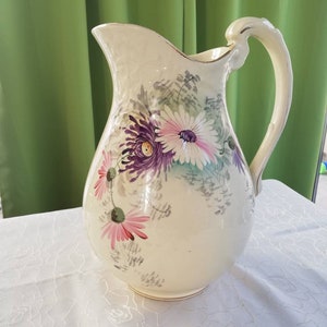May include: Cream ceramic pitcher with a floral design. The pitcher has a wide spout, a curved handle, and is decorated with purple, pink, and white flowers. The rim is trimmed with gold. The pitcher is on a white tablecloth.