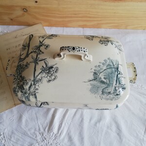 Op de afbeelding: Een witte keramische serveerschaal met een zwart en groen bloemenmotief. De schaal heeft een deksel met een handvat en een zwart en wit bloemenmotief.