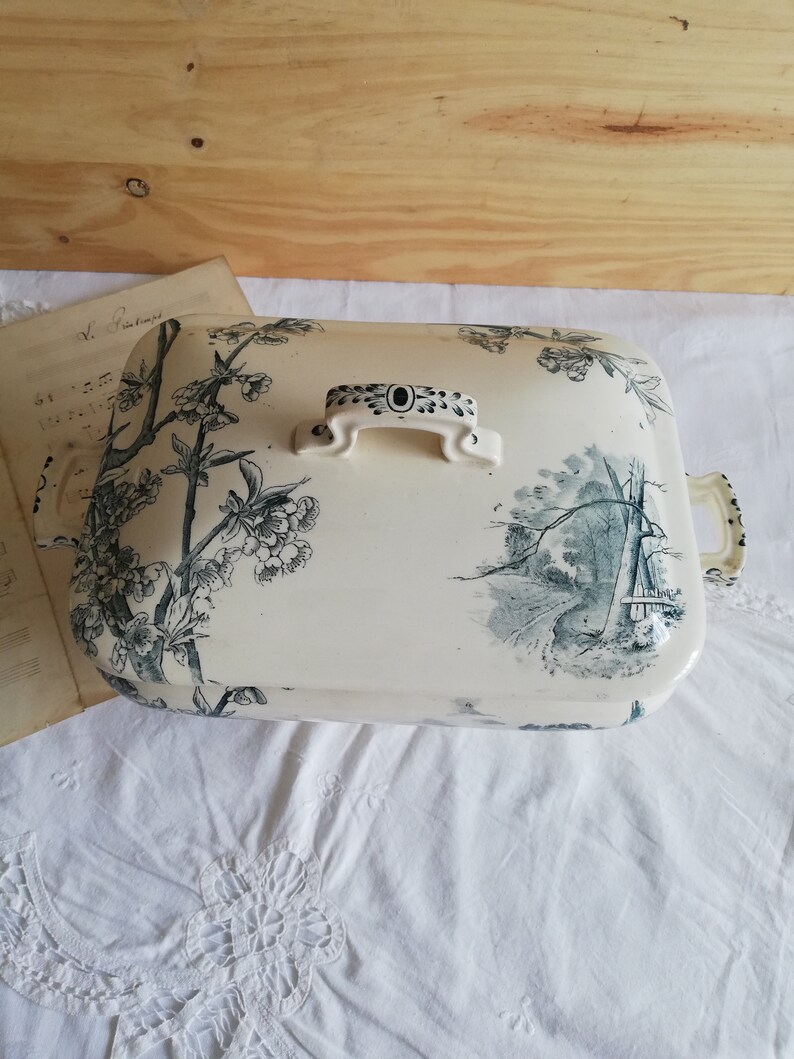 Op de afbeelding: Een witte keramische serveerschaal met een zwart en groen bloemenmotief. De schaal heeft een deksel met een handvat en een zwart en wit bloemenmotief.
