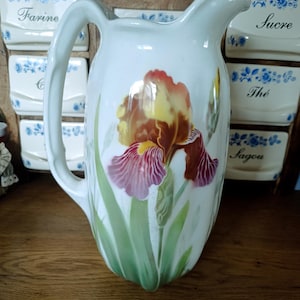 Op de afbeelding: Een witte keramische kan met een grote, handgeschilderde irissbloem in tinten geel, oranje en dieppaars. Het bloemmotief bevat lange groene stengels en bladeren die vanaf de basis omhoog groeien. De kan heeft een gebogen handvat en een schenktuit.