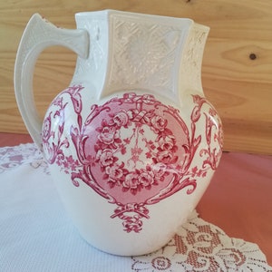 Op de afbeelding: Witte keramische kan met een roze bloemenpatroon. De kan heeft een vierkante bovenkant en een rond lichaam. Het bloemenpatroon heeft roze rozen en bladeren.