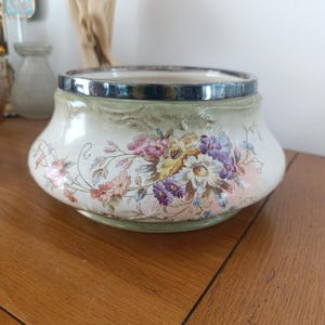 May include: A white ceramic bowl with a silver rim and a floral design. The bowl has a green background with pink, yellow, and purple flowers.