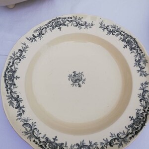May include: A white ceramic plate with a black floral design around the edge. The plate has a scalloped edge and a small floral design in the center.