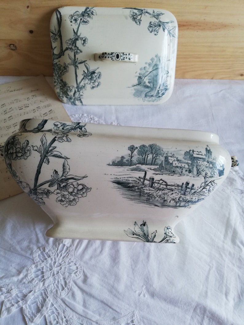 Op de afbeelding: Een witte keramische terrine met een blauw-wit bloemenpatroon. De terrine heeft een deksel met een bijpassend patroon en een handvat. De terrine staat op een wit tafelkleed.