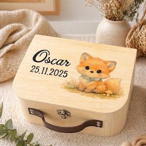 Pode incluir: Caixa de madeira clara com alça e fecho. Apresenta uma ilustração de raposa de desenho animado e o nome "Oscar" com a data "25.11.2025" impressos na tampa. A raposa é laranja com um laço azul. Ideal para guardar.