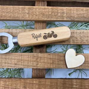 May include: A bottle opener with a light-colored wooden handle featuring a laser-engraved motorcycle graphic and the name "Rafa&euml;l" in black script. A small, plain wooden heart shape is positioned next to the bottle opener on a slatted surface.