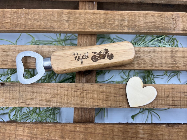 May include: A bottle opener with a light-colored wooden handle featuring a laser-engraved motorcycle graphic and the name "Rafa&euml;l" in black script. A small, plain wooden heart shape is positioned next to the bottle opener on a slatted surface.