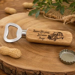May include: A bottle opener with a light brown wooden handle featuring a laser-engraved illustration of a vintage car and the French text "Bonne f&ecirc;te papi Je t'aime Hugo". The metal opener head is silver-colored and attached to the wooden handle.