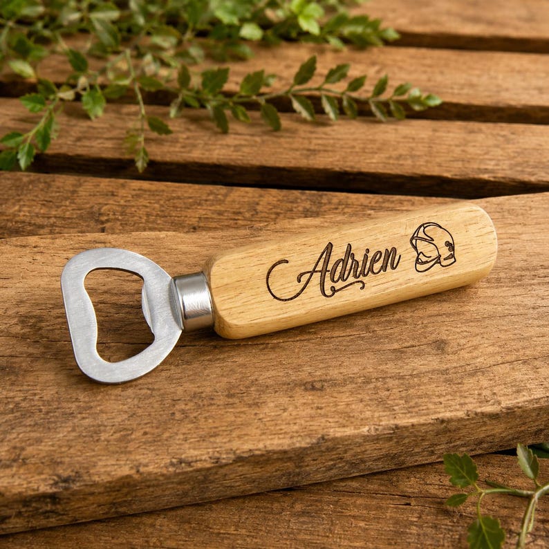 May include: A bottle opener with a light-colored wooden handle featuring the engraved name Adrien in a cursive script and a small graphic of a firefighter helmet. The metal opener head is silver-toned and attached to the wooden handle.