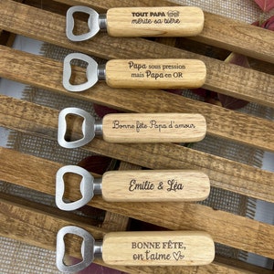 May include: Five bottle openers with light-colored wooden handles and silver metal heads. Each handle features unique laser-engraved text in French, including phrases like "TOUT PAPA m&eacute;rite sa bi&egrave;re," "Papa sous pression," and "Bonne f&ecirc;te, on t'aime."
