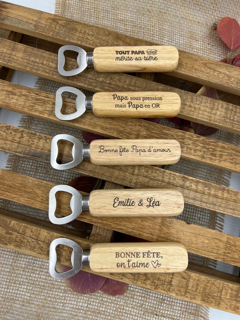 May include: Five bottle openers with light-colored wooden handles and silver metal heads. Each handle features unique laser-engraved text in French, including phrases like "TOUT PAPA m&eacute;rite sa bi&egrave;re," "Papa sous pression," and "Bonne f&ecirc;te, on t'aime."