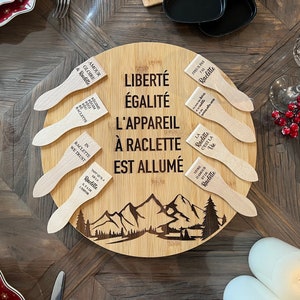 Peut inclure: Un plateau de service en bois avec une scène de montagne et le texte "Liberté Égalité L'appareil à Raclette est allumé". Le plateau comporte six spatules en bois avec diverses citations sur la raclette, notamment "Amour & Gloire & Raclette", "In Raclette We Trust", et "Tant qu'il a de la Raclette, il y a de l'espoir".