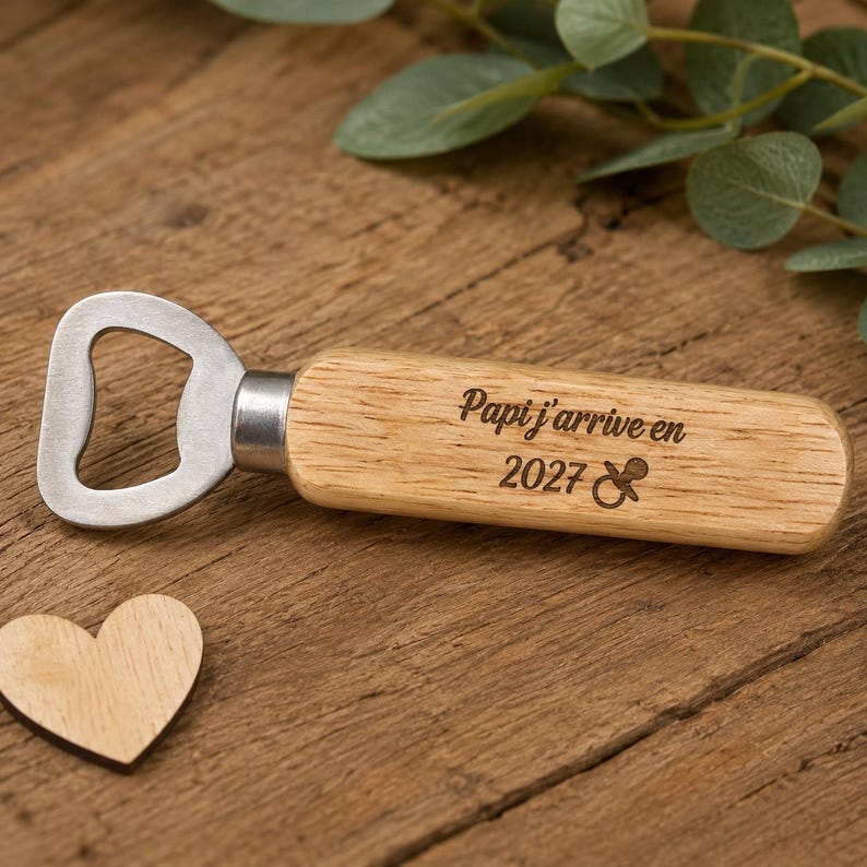 May include: A bottle opener with a light brown wooden handle featuring the laser-engraved text Papi j'arrive en 2027 and a small pacifier icon. The metal opener head is silver-colored and attached to the handle with a circular metal ferrule.