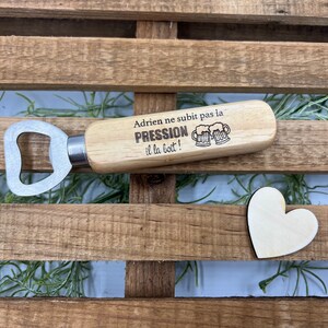 May include: A wooden bottle opener with a metal head and a handle engraved with the text "Adrien ne subit pas la PRESSION il la boit!" alongside an illustration of two beer mugs. A small, plain wooden heart shape is positioned nearby.