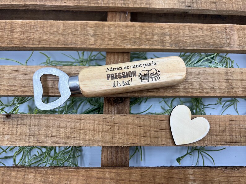 May include: A wooden bottle opener with a metal head and a handle engraved with the text "Adrien ne subit pas la PRESSION il la boit!" alongside an illustration of two beer mugs. A small, plain wooden heart shape is positioned nearby.