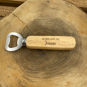 May include: A bottle opener with a light-colored wooden handle featuring the engraved text MARRAINE DE Jeanne. The metal opener head is silver-toned and attached to the handle with a polished metal ferrule, creating a functional tool.
