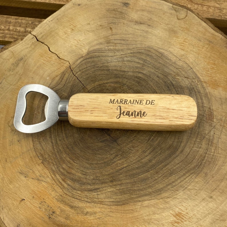 May include: A bottle opener with a light-colored wooden handle featuring the engraved text MARRAINE DE Jeanne. The metal opener head is silver-toned and attached to the handle with a polished metal ferrule, creating a functional tool.