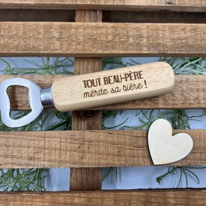 May include: A bottle opener with a light-colored wooden handle featuring the engraved French text "TOUT BEAU-P&Egrave;RE m&eacute;rite sa bi&egrave;re !" and a silver-toned metal head, accompanied by a small, flat, light-colored wooden heart shape.