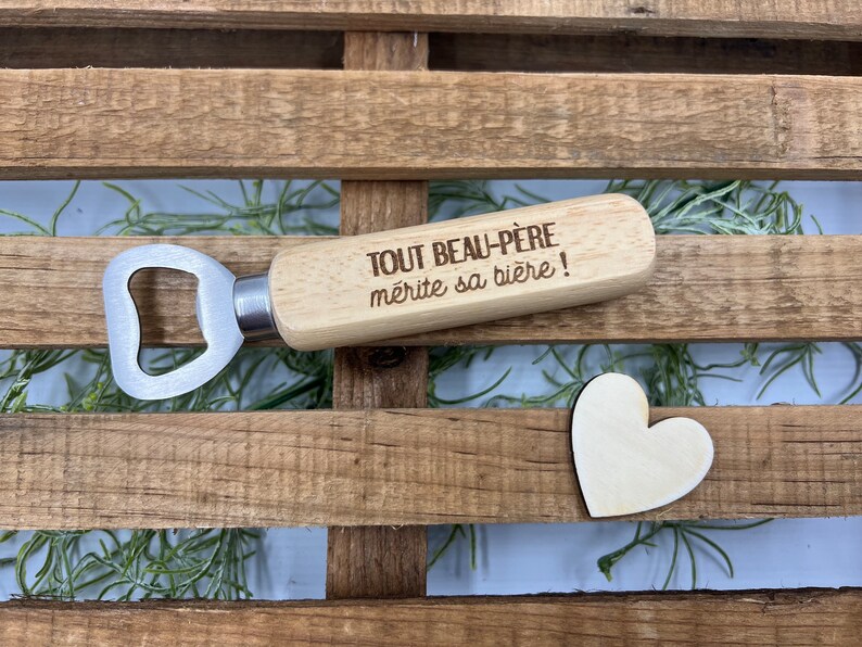 May include: A bottle opener with a light-colored wooden handle featuring the engraved French text "TOUT BEAU-P&Egrave;RE m&eacute;rite sa bi&egrave;re !" and a silver-toned metal head, accompanied by a small, flat, light-colored wooden heart shape.