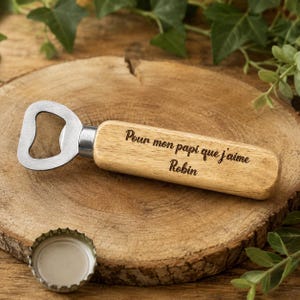 May include: A bottle opener with a light brown wooden handle featuring the engraved text Pour mon papi que j'aime and the name Robin. The opener has a silver metal head and is positioned next to a silver metal bottle cap on a wooden surface.