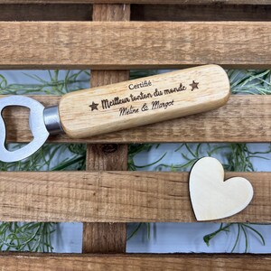 May include: A wooden bottle opener with a silver metal head. The handle is engraved with the text "Certifi&eacute; Meilleur tonton du monde" flanked by two stars, followed by the names "M&eacute;line & Margot". A small wooden heart shape is placed nearby.