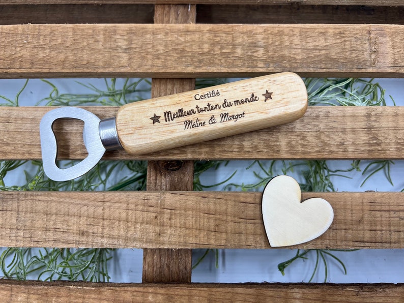 May include: A wooden bottle opener with a silver metal head. The handle is engraved with the text "Certifi&eacute; Meilleur tonton du monde" flanked by two stars, followed by the names "M&eacute;line & Margot". A small wooden heart shape is placed nearby.