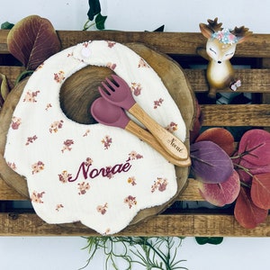 May include: A white bib with a floral pattern and the name "Novae" embroidered in pink. The bib is on a wooden surface with a pair of wooden utensils with pink silicone tips.