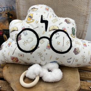 Peut inclure: Un oreiller en forme de nuage blanc avec un contour noir des lunettes de Harry Potter et un éclair. L'oreiller est décoré sur le thème d'Harry Potter, avec des étoiles, des baguettes magiques et des livres. Un jouet en peluche blanc avec un anneau en bois se trouve devant l'oreiller.