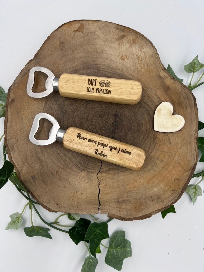 May include: Two bottle openers with light-colored wooden handles and metal heads. The top opener is engraved with the text PAPI SOUS PRESSION and a beer mug icon. The bottom opener is engraved with the text Pour mon papi que j'aime Robin.