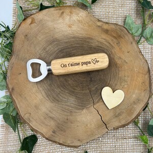 May include: A bottle opener with a light-colored wooden handle featuring the engraved text On t'aime papa and a small heart symbol, accompanied by a separate small wooden heart shape, both resting on a natural wood slice surface.